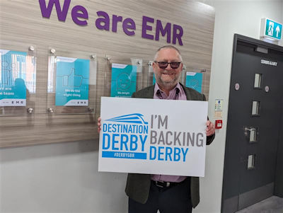 Man holding an 'I'm backing Derby' sign in front of a 'We are EMR' sign